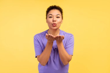 Gülümseyen Afrikalı Amerikalı kadının portresi, el ele tutuşan, öpüşen, sarı arka planda izole bir şekilde duran kameraya bakan. Pozitif yaşam tarzı kavramı