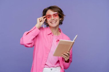 Şık gözlükler takan kendine güvenen üniversite öğrencisi elinde açık bir kitapla menekşe arka planda, eğitim konseptinde izole edilmiş. Güzel bir portre mutlu bir kadın, çalışan, kameraya bakan serbest çalışan. 