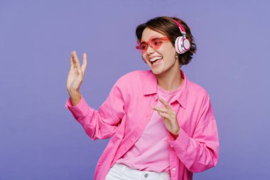 Güneş gözlüklü, kulaklık takan, müzik dinleyen, menekşe arka planda izole edilmiş, dans eden, eğlenen güzel bir kadın. Mutlu yaşam tarzı konsepti 