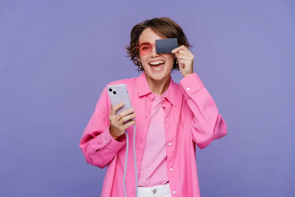 微笑的现代女人拿着手机 用信用卡盖住眼睛 网上购物 隔离在紫罗兰的背景下 移动银行的概念 — 图库照片