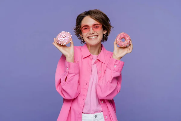 Portrait of excited woman holding pink donuts, looking at camera isolated on violet background. Concept of food, dessert