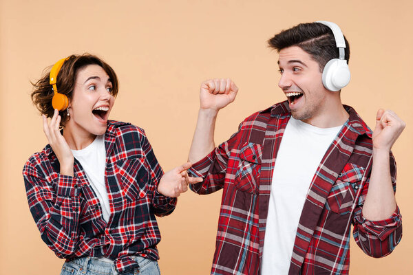 Young overjoyed couple wearing casual clothing listening music, dancing, singing, having fun isolated on beige background. Technology, positive emotions concept 