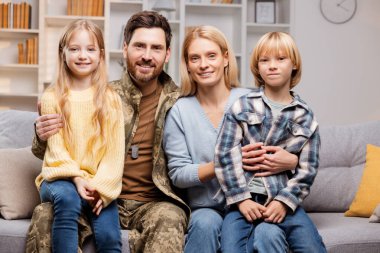 Bir kahramanın eve dönüşü: Baba Ukrayna askeri kıyafetleriyle giyinmiş, neşeli ailesiyle rahat kanepelerinde şefkatli kucaklaşmalar yapıyor, sade bir ortamda mutlu bir aile portresi sergiliyor.