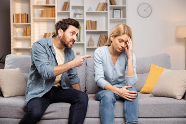 Oturma odasında, kocası bağırıp, parmakla gösterip, aile dinamiklerinde desteğe olan umutsuz ihtiyacını gösterirken, bir kadın umutsuzluk içinde başını tutar.