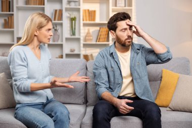 Help for families under strain. A woman, enraged, confronts her husband in their living space, as he exhibits signs of a headache by holding his forehead clipart