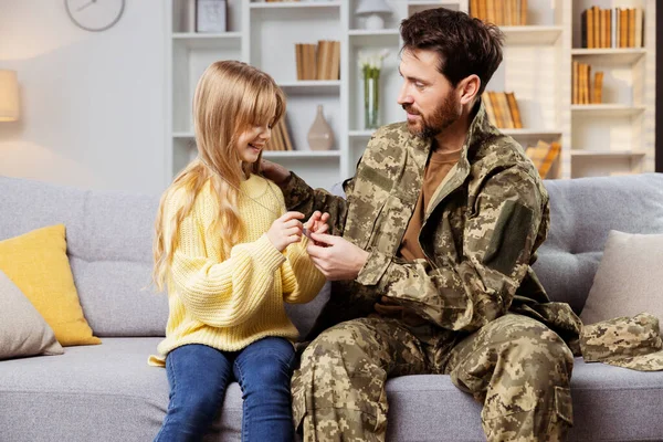 Back from duty, a soldier in Ukrainian gear lounges at home with his cheerful daughter, entrusting her with his dog tag, a sign of his enduring presence
