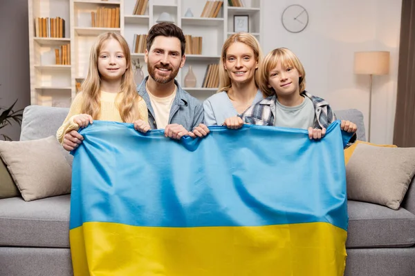 In a warm home setting, a devoted Ukrainian family, including mom, dad, and two kids, sits closely on a sofa, united in displaying the Ukrainian flag, symbolizing their strong patriotic bond