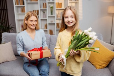 Evde sıcak bir Anneler Günü sahnesi, elinde buketi tutan ve kameraya bakan küçük bir kız, kanepede rahat bir şekilde oturan annesi, hediye kutusunu alır ve çocuğuna bakar.