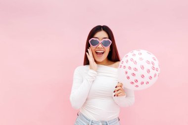 Gözlüklü Asyalı kadının balon tutmasına ve pembe arka planda poz verirken kameraya bakmasına çok şaşırdım. Sevgililer Günü Kutlama Konsepti