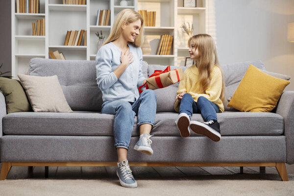 Празднуя День матери в помещении, маленькая девочка удивляет свою мать подарочной коробкой на удобном диване. Мать, ошеломленная радостью, с трепетом кладет руку на грудь