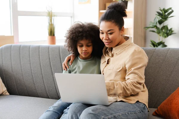 A heartwarming photo of an African American mom and her little girl, sitting closely on a couch with a laptop, sharing a joyful moment together. Happy family at home concept