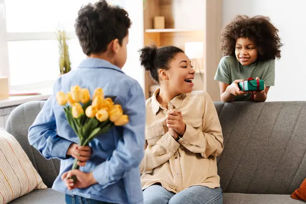 Heartwarming Mother\'s Day at home, capturing amazed African American mother, receiving presents from her kids. Her daughter presents gift box, and her son conceals floral bouquet, ready to surprise