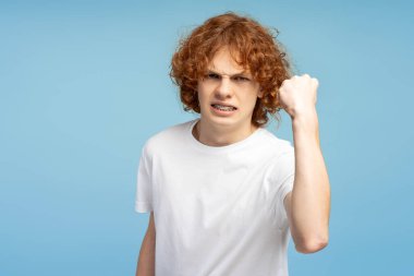 Kızgın kıvırcık çocuk yumruğunu sıktı ve mavi arka planda tek başına savaşmaya hazır. Saldırgan öğrenci saldırmaya hazırlanıyor, stüdyo çekimi. 