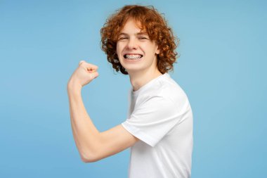 Kaslarını gösteren şık beyaz tişört giyen güçlü heyecanlı genç mavi arka planda izole kameraya bakıyor. Etkin yaşam tarzını kavra 