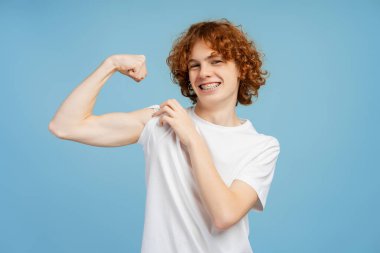 Diş teli takan, bisepsi işaret eden, mavi arka planda izole edilmiş kameraya bakan yakışıklı ve güçlü bir çocuk. Reklam kavramı 