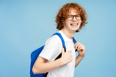 Sırt çantasında diş telleri olan zeki gülümseyen üniversite öğrencisi mavi arka planda izole edilmiş kameraya bakıyor. Eğitim kavramı, kopyalama alanı 