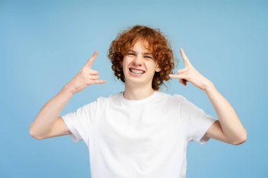 Tişört giyen, kaya işareti gösteren, mavi arka planda izole edilmiş kameraya bakan neşeli, neşeli genç bir adam. Pozitif yaşam tarzı, duygu konsepti 