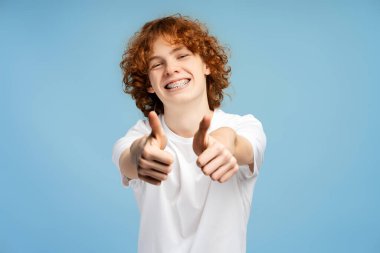 Gülümseyen kıvırcık saçlı gencin portresi, beyaz tişört giyen hippi mavi arka planda izole edilmiş havalı bir jestle kameraya bakıyor. Fotoğraflar için mutlu bir model, stüdyo çekimi. 