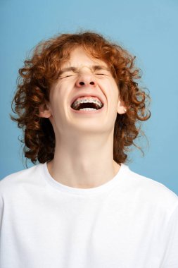 Gülen çocuğun dikey portresi, dişlerinde diş teli olan, mavi arka planda eğlenen kıvırcık kızıl saçlı genç.