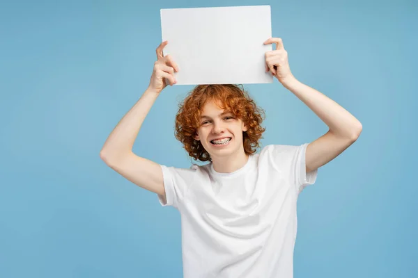 青い背景に隔離された空のプラカードを保持するブレースを持つ幸せなカーリー少年 モックアップ カメラを見ているポスターとスマートな笑顔の学生の肖像 — ストック写真