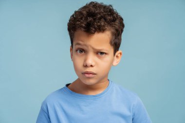 Ciddi, çekici, Afro-Amerikalı, sıradan tişörtler giyen, mavi arka planda izole edilmiş kameraya bakan yakışıklı bir çocuk. Eğitim kavramı, reklam