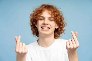 Çekici kıvırcık saçlı genç bir kızın portre resmi. Diş telleriyle gülümseyen ve Kore 'de kameraya aşk jesti yapan, mavi bir stüdyo arka planında çekilmiş.