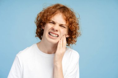 Kıvırcık kızıl saçları ve diş telleri olan, yanağına bastıran, diş ağrısını vurgulayan, mavi arka planda izole edilmiş sıkıntılı bir gencin portresi. Diş sağlığı konsepti