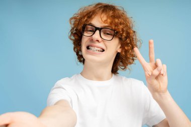 Kıvırcık kızıl saçlı, diş telli, gözlüklü ve mavi arka planda barış işareti yapan gülümseyen bir gencin selfie 'si.
