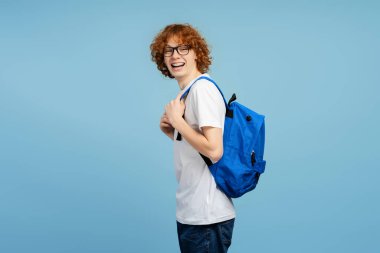 Duygusal, kıvırcık saçlı, gözlüklü ve sırt çantalı bir gencin görüntüsü. Profilde yer almış, gülüyor ve kameraya bakıyor. Mavi arka planda izole edilmiş. Öğrenim meraklısı kavramı