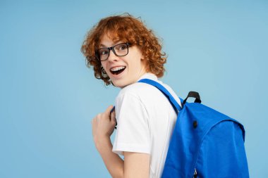 Heyecanlı kızıl saçlı, kıvırcık saçlı ve ortodontik telli bir gencin fotoğrafı. Gözlük ve sırt çantasıyla donatılmış, omzunun üstünden bakan, mavi bir arka planda izole edilmiş.