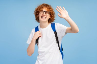 Kırmızı, mutlu, kıvırcık saçlı, diş teli takan, göz ve sırt çantasıyla giyinmiş, kameraya el sallayan, mavi arka planda izole edilmiş genç bir erkek. Bilimsel inek konseptini gözler önüne seriyor.