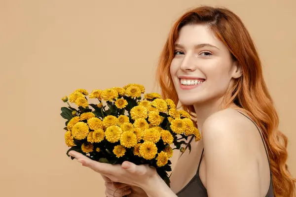 Portrait beautiful smiling cute woman holding bouquet of yellow chrysanthemums flowers looking at camera isolated on beige background. Natural beauty, spring, International women\'s day concept