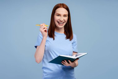 Mavi tişörtlü, kendine güveni tam bir üniversite öğrencisi elinde mavi arka planda izole bir kitap tutuyor. Eğitim kavramı. Mutlu kadın portresi, işçi, kameraya bakan serbest çalışan. 