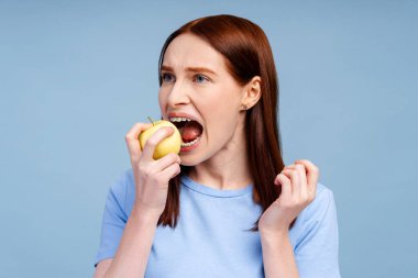 Mavi tişörtlü, yeşil elmalı ve mavi arka plan stüdyo portresini ısıran kızıl saçlı, gülümseyen kadın. Sağlık prosedürleri kavramı