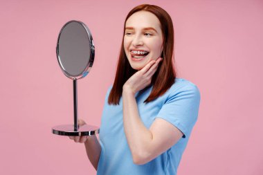 Çekici kızıl kadın aynaya bakıyor, göz kırpıyor, pembe arka planda izole edilmiş dişleriyle tatmin oluyor. Diş bakımı ve sağlık kavramı