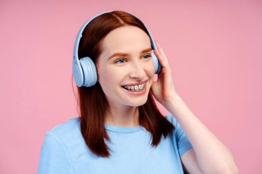Diş telleriyle gülümseyen çekici kızıl kadının portresi pembe arka planda izole edilmiş kablosuz kulaklıkla müzik dinliyor.