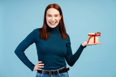 Hediye kutusu tutan kadının portresi, bayramı kutlamak için doğum günü hediyesi, kameraya bakmak. Mavi arka planda kapalı stüdyo çekimi 