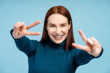 Mavi balıkçı yaka giyen neşeli kızıl kadın mavi arka planda izole edilmiş kameraya bakarken zafer işareti gösteriyor. Reklam kavramı