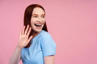 Sosyal kızıl saçlı, diş teli takan mutlu güzel bir kadın, tişört giyiyor, selam veriyor ve yüzünü çeviriyor, pembe bir arka planda soyutlanmış.