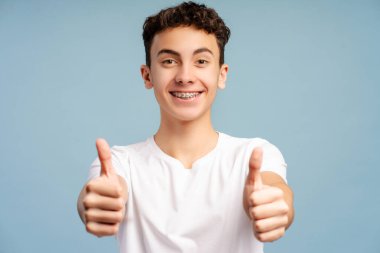 Gülümseyen yakışıklı bir çocuğun portresi, diş teli takan, şık beyaz tişört giyen, mavi arka planda izole kameraya bakan 15 yaşında bir genç. Reklam kavramı 