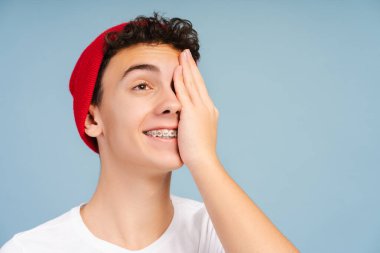 Gülümseyen kıvırcık saçlı diş teli takmış bir çocuğun portresi mavi arka planda izole edilmiş bir gözünü kaçırıyor. Sağlık hizmeti, ortodontik konsept 