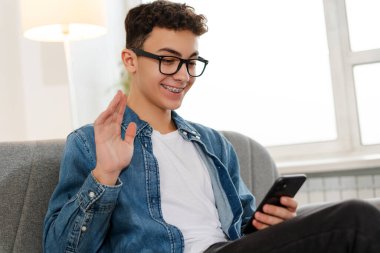 Gülümseyen çocuk, diş telleriyle cep telefonu tutan genç, internetten iletişim kuran, elini selamlayan, rahat bir koltukta video görüşmesi yapan, evde dinlenen. Teknoloji, sosyal medya kavramı 