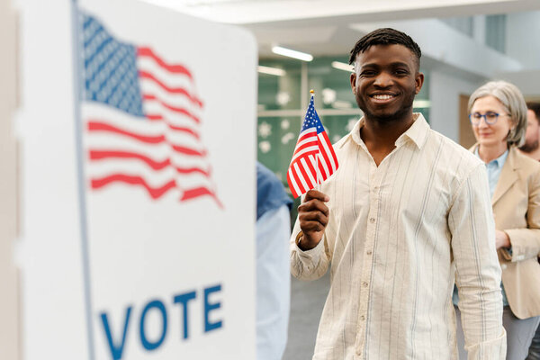 Portrait of smiling African American man holding American flag vote at polling station. Democracy, freedom, United stated election day concept 