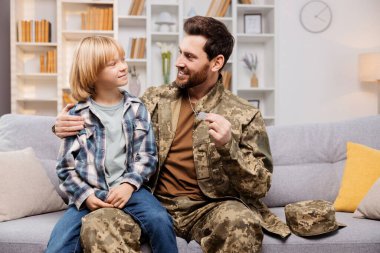 İçeride mutlu bir askeri aile, üniformalı gülümseyen bir baba, gururlu oğlunu tutuyor ve kimlik kartını gösteriyor. Kanepede oturuyorlar, kucaklaşıyorlar.