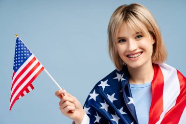 Gülümseyen, sarı saçlı, elinde Amerikan bayrağı olan genç ve güzel bir kadın kameraya bakıyor. Mavi arka planda tek başına duran kadın destekçi. Amerikan bağımsızlık günü konsepti 
