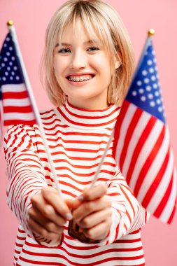 Amerikan bayraklarıyla gülümseyen kadının dikey görüntüsü, kameraya bakıyor, pembe arka planda izole bir şekilde poz veriyor. Seçim sonuçlarını bekleyen neşeli bayan destekçi. Cumhurbaşkanlığı seçimleri 