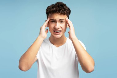 Yorgun, üzgün, telleri kafasına değen, migren, depresyon geçiren, kameraya bakan bir çocuk. Üzgün genç, mavi arka planda izole edilmiş baş ağrısı çekiyor. Sağlık hizmeti kavramı