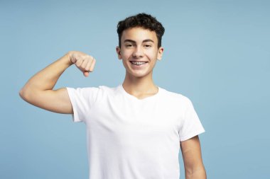 Mavi arka planda izole edilmiş kameraya bakarken kaslarını gösteren şık beyaz tişört giyen güçlü bir gencin portresi. Reklam kavramı