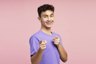 Diş teli takan, parmakla gösterip kameraya bakan gülümseyen yakışıklı çocuğun portresi. Pembe arka planda şık kıyafetler giyen çekici bir genç. Alışveriş konsepti, reklam