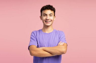 Dişlerinde diş teli olan, kolları çapraz mor tişörtlü ve pembe arka planda izole edilmiş kameraya bakan kendine güvenen bir çocuğun portresi. Reklam kavramı